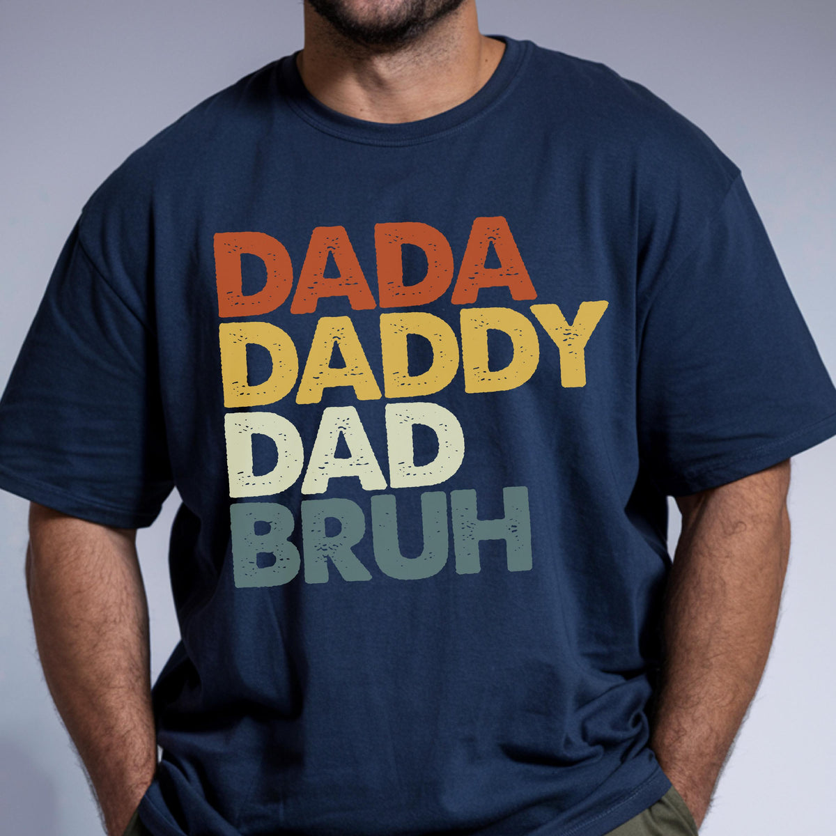 May include: Navy blue t-shirt with the words "DADA DADDY DAD BRUH" in orange, yellow, off-white, and green. The text is stacked vertically, creating a retro, vintage look. A casual, comfortable tee.