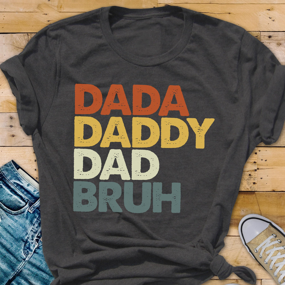 May include: Dark gray t-shirt with the words "DADA DADDY DAD BRUH" in orange, yellow, cream, and teal lettering. The shirt is a casual style, suitable for everyday wear.