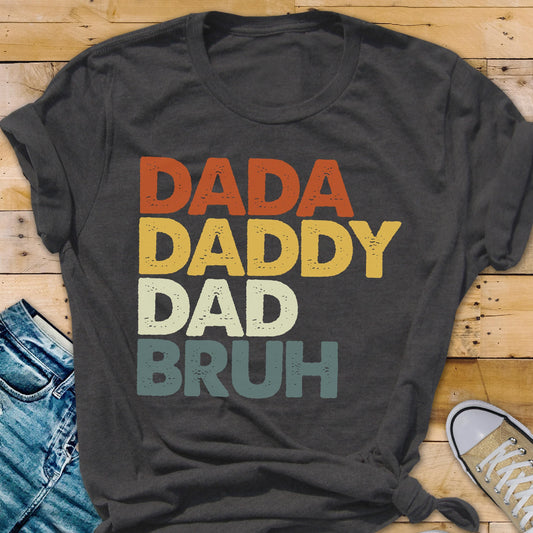 May include: Dark gray t-shirt with the words "DADA DADDY DAD BRUH" in orange, yellow, cream, and teal lettering. The shirt is a casual style, suitable for everyday wear.