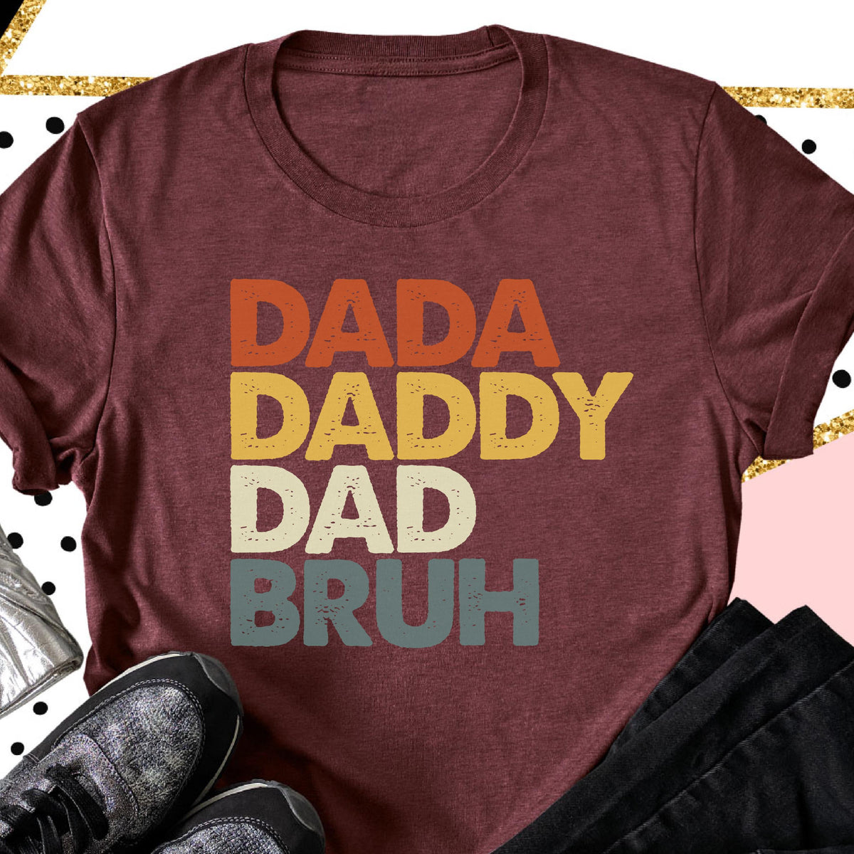 May include: Burgundy t-shirt with the words "DADA DADDY DAD BRUH" in orange, yellow, white, and blue-green block letters. The shirt is folded, with a pair of gray and black sneakers and black pants visible in the foreground.