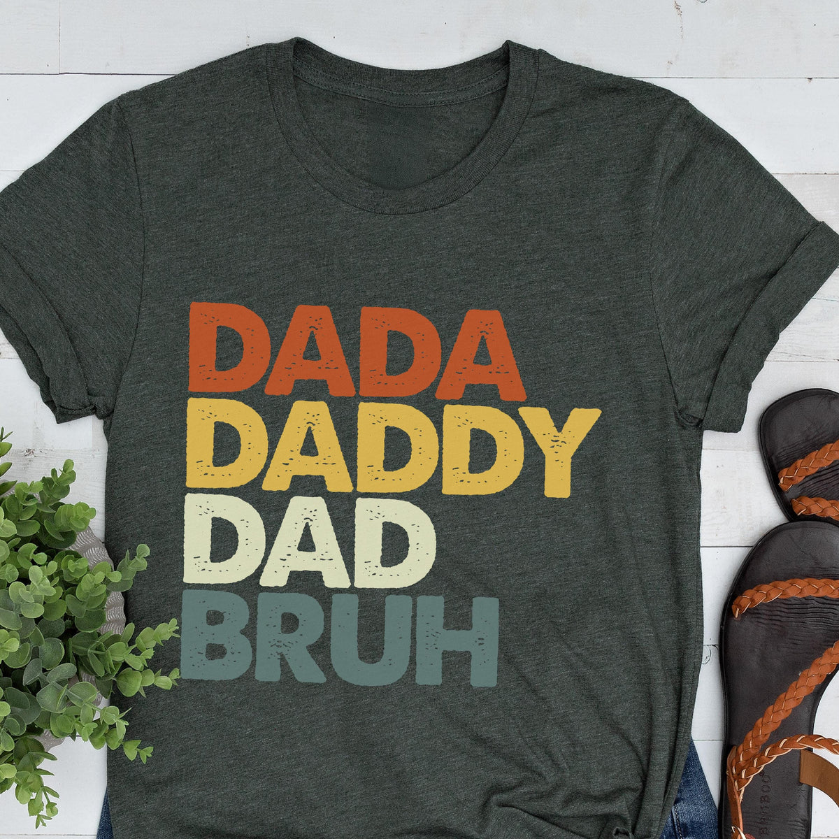 May include: Dark gray t-shirt with the words "DADA DADDY DAD BRUH" in orange, yellow, white, and teal. The shirt has a crew neck and short sleeves. The text is in a retro, layered font.
