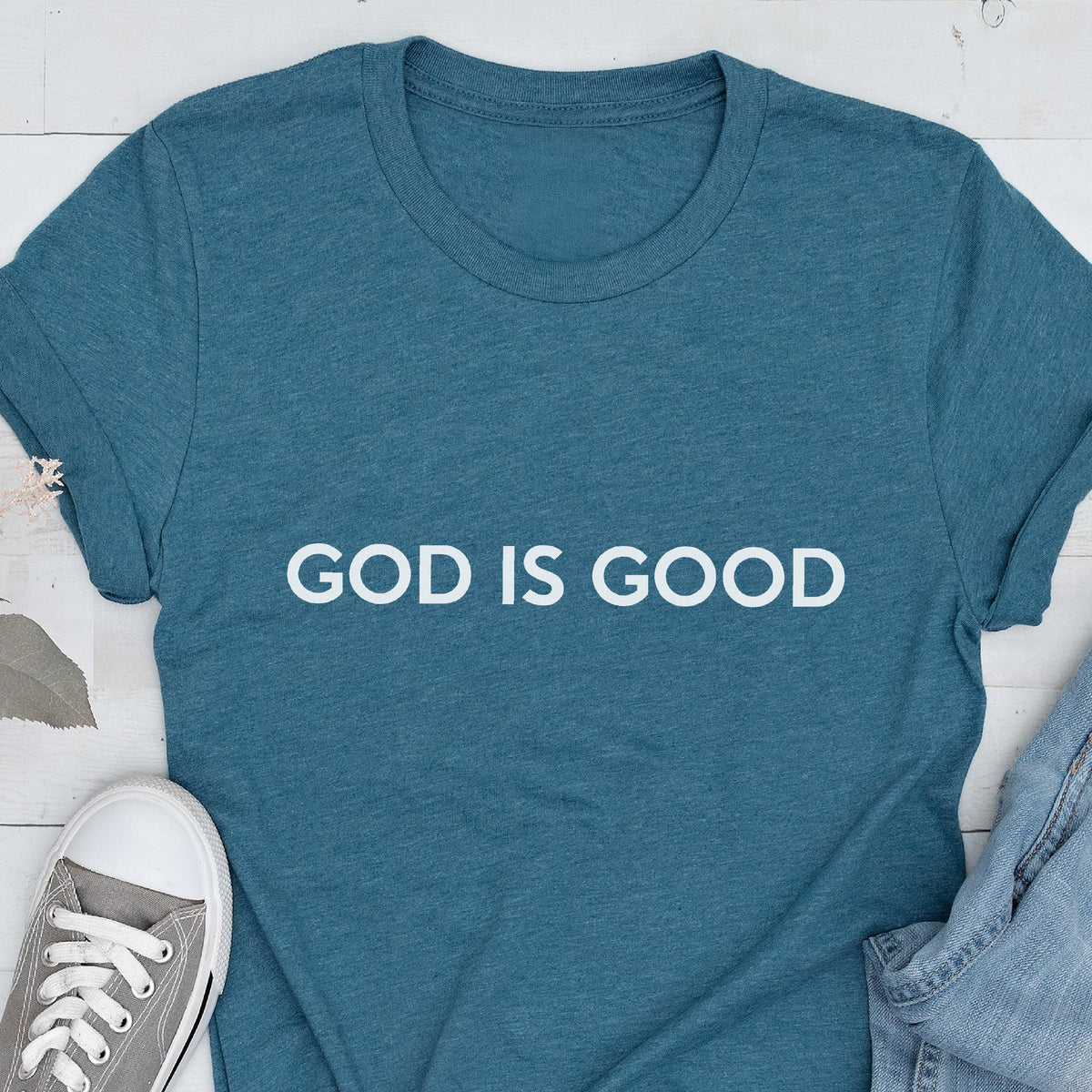 May include: Teal t-shirt with the white text "GOD IS GOOD". The shirt is made of a soft material and has short sleeves. A gray sneaker and denim jeans are partially visible in the image.