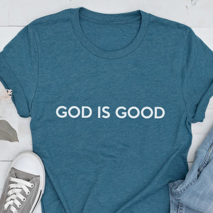 May include: Teal t-shirt with the white text "GOD IS GOOD". The shirt is made of a soft material and has short sleeves. A gray sneaker and denim jeans are partially visible in the image.