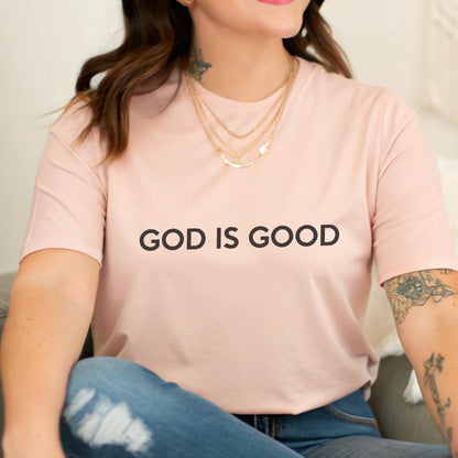 May include: A light pink t-shirt with the text "GOD IS GOOD" in black. The shirt is made of a soft material and is being worn by a person. The person is also wearing gold necklaces and blue jeans.