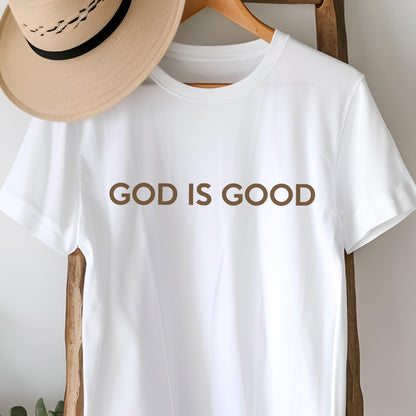 May include: A white t-shirt with the phrase "GOD IS GOOD" printed in brown. A tan hat with a black band is visible in the background, hanging on a wooden hanger.