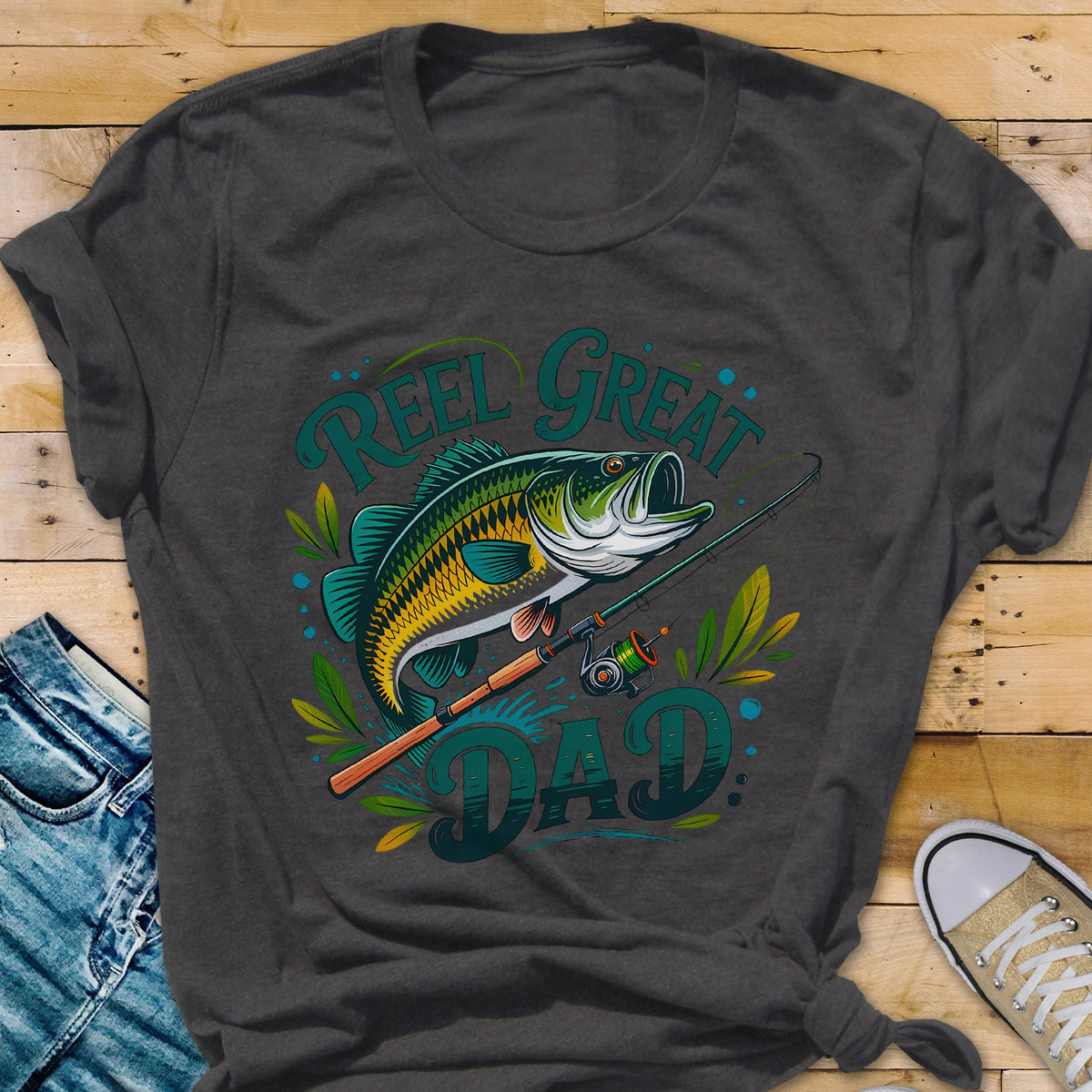 May include: Dark gray t-shirt featuring a colorful illustration of a fish, fishing rod, and the words "REEL GREAT DAD" in teal. The fish is green, yellow, and white. The fishing rod is brown and green. The shirt is a crew neck.