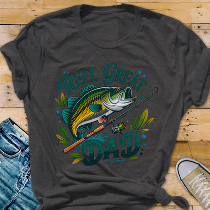 May include: Dark gray t-shirt featuring a colorful illustration of a fish, fishing rod, and the words "REEL GREAT DAD" in teal. The fish is green, yellow, and white. The fishing rod is brown and green. The shirt is a crew neck.