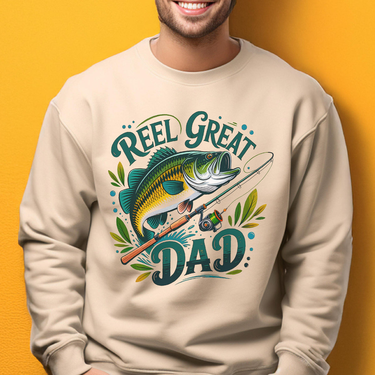 May include: Beige sweatshirt with a graphic featuring a green and yellow fish, fishing rod, and the text "Reel Great Dad" in teal. The design includes decorative leaves and dots, perfect for fishing enthusiasts.