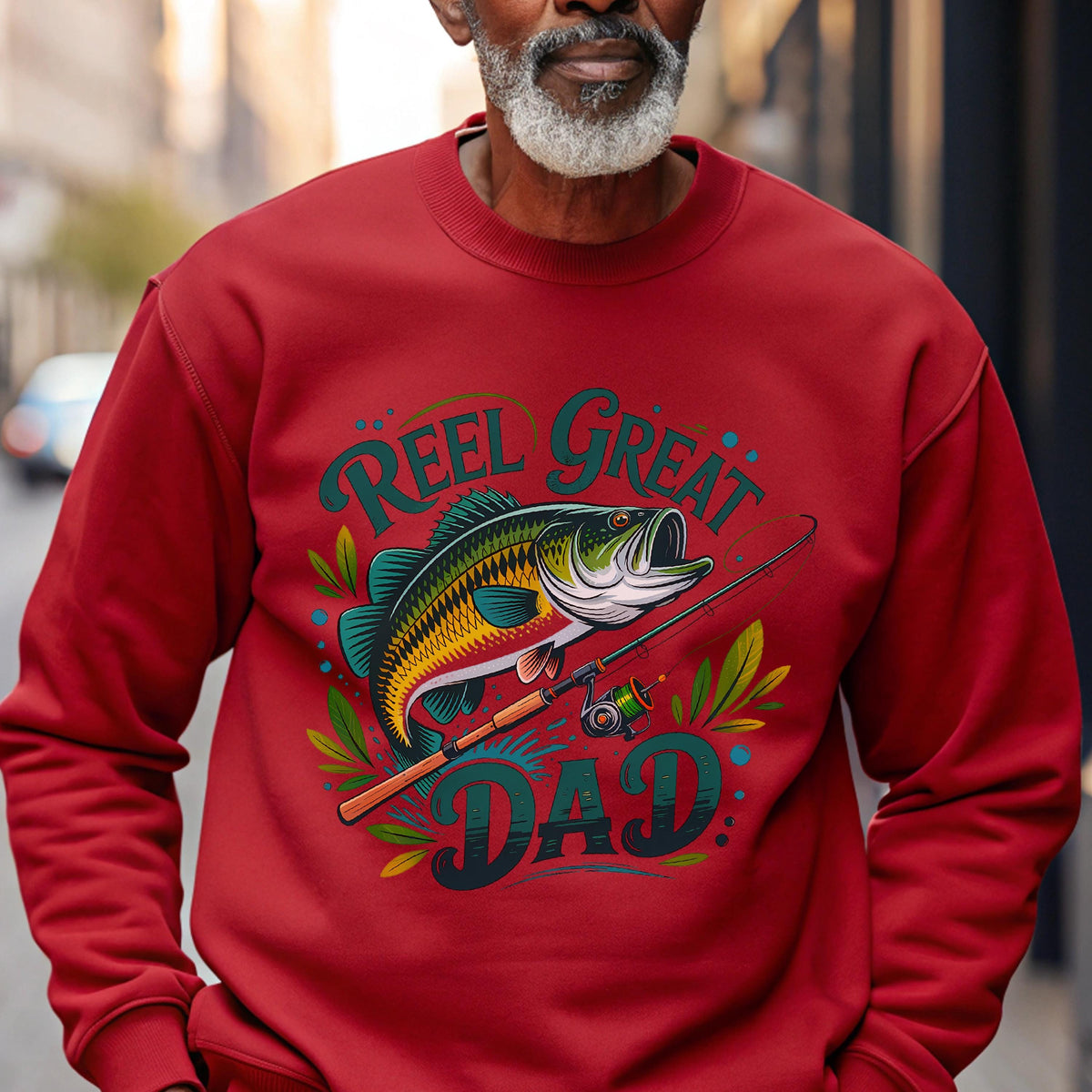 May include: Red sweatshirt with a graphic of a fish, fishing rod, and the words "REEL GREAT DAD" in teal and yellow. The fish is detailed with yellow, green, and black scales. A fishing-themed apparel item.