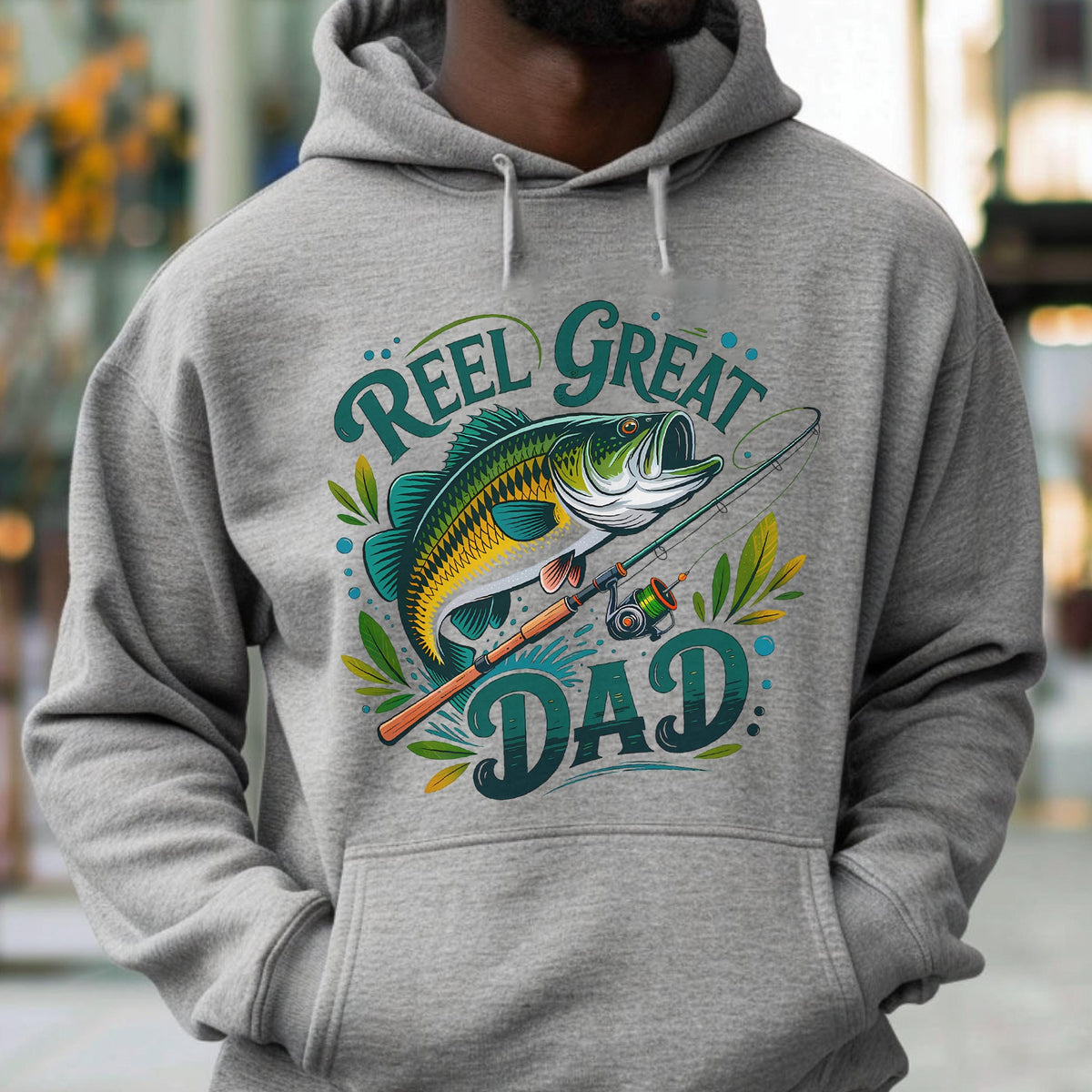 May include: A gray hoodie featuring a graphic of a fish, fishing rod, and the words "Reel Great Dad" in teal and white. The design includes leaves and dots, creating a fishing-themed design.