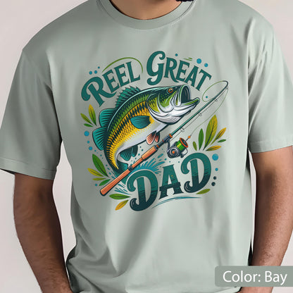 May include: A light green t-shirt with a colorful graphic of a fish and fishing rod. The text "REEL GREAT DAD" is in a teal script font. The shirt is a solid color and has a crew neck.