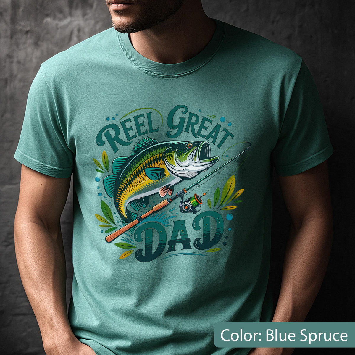 May include: Blue spruce t-shirt with a graphic of a fish, fishing rod, and the words "Reel Great Dad." The design features a detailed illustration of a fish with yellow and green scales, a fishing rod, and decorative leaves.