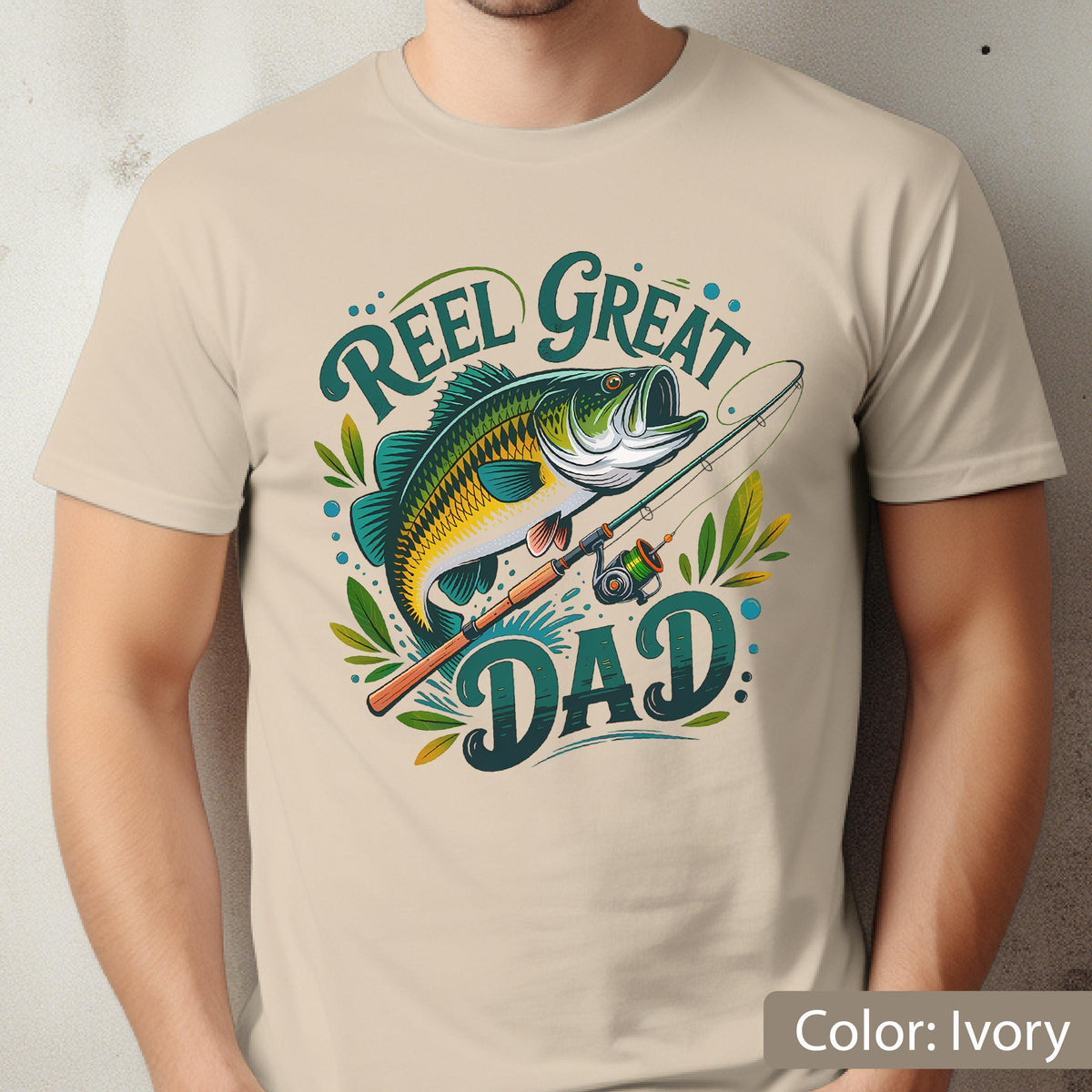 May include: Ivory-colored t-shirt featuring a colorful graphic of a fish, fishing rod, and the words "Reel Great Dad." The design includes shades of green, blue, yellow, and brown. The shirt is made of a soft material.