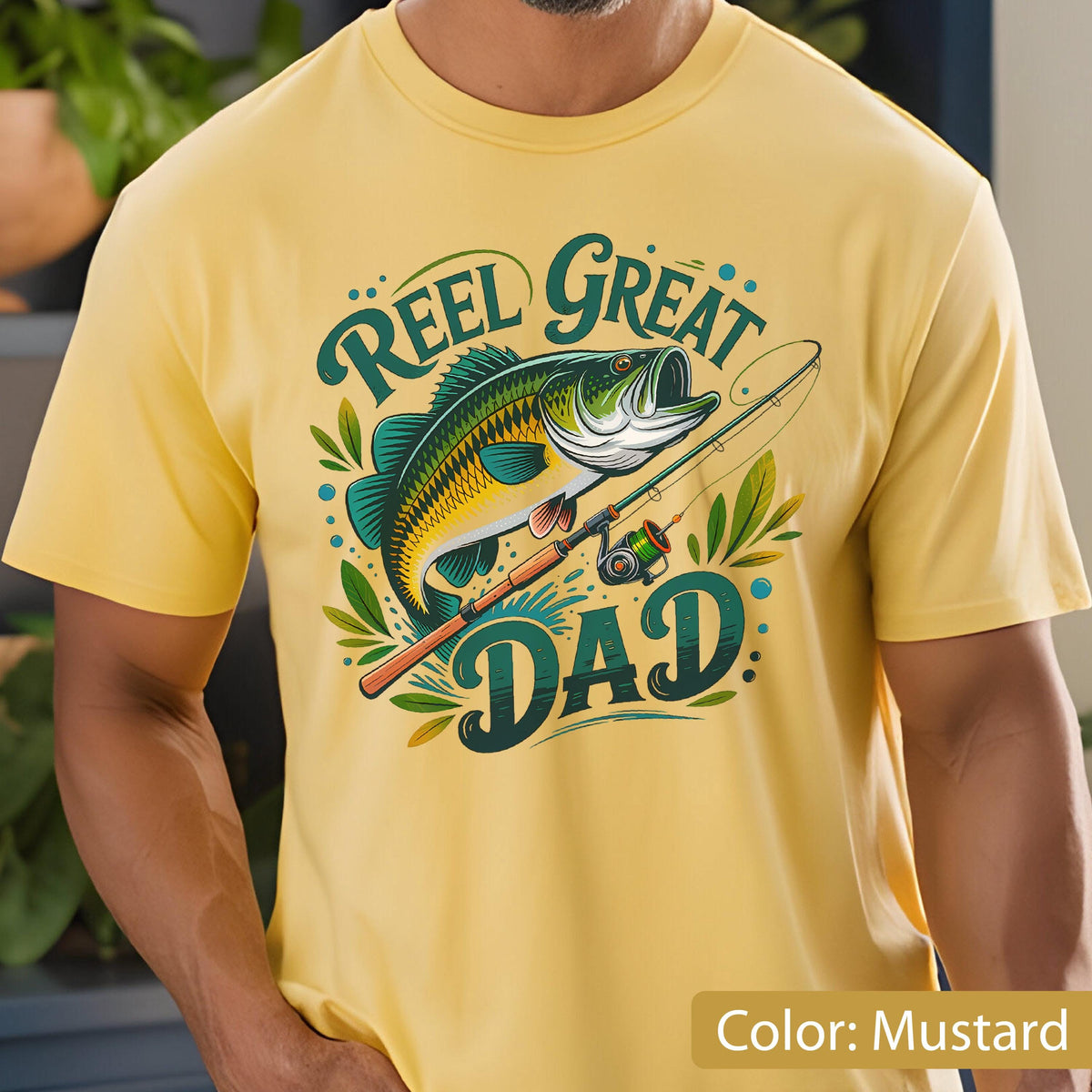 May include: Mustard yellow t-shirt featuring a graphic of a fish, fishing rod, and the words "Reel Great Dad" in teal and green. The design includes leaves and dots. The shirt is short-sleeved and made of a soft material.