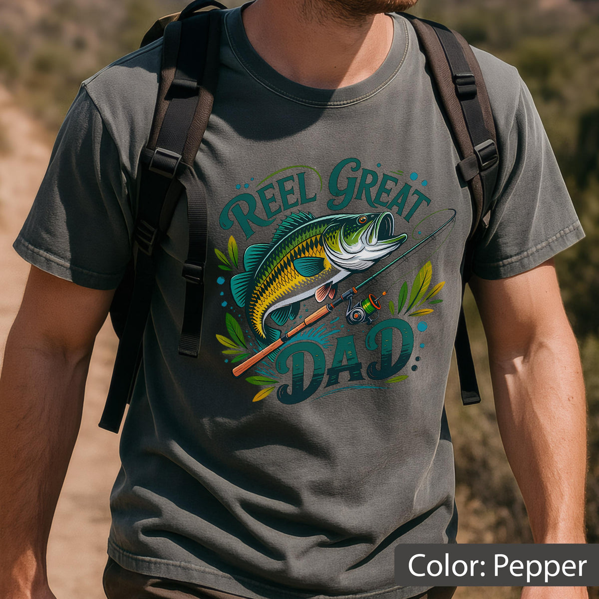 May include: Pepper-colored t-shirt featuring a graphic of a largemouth bass, fishing rod, and the text "Reel Great Dad." The design includes shades of green, yellow, and blue. The shirt is a short-sleeved crew neck.