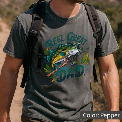 May include: Pepper-colored t-shirt featuring a graphic of a largemouth bass, fishing rod, and the text "Reel Great Dad." The design includes shades of green, yellow, and blue. The shirt is a short-sleeved crew neck.
