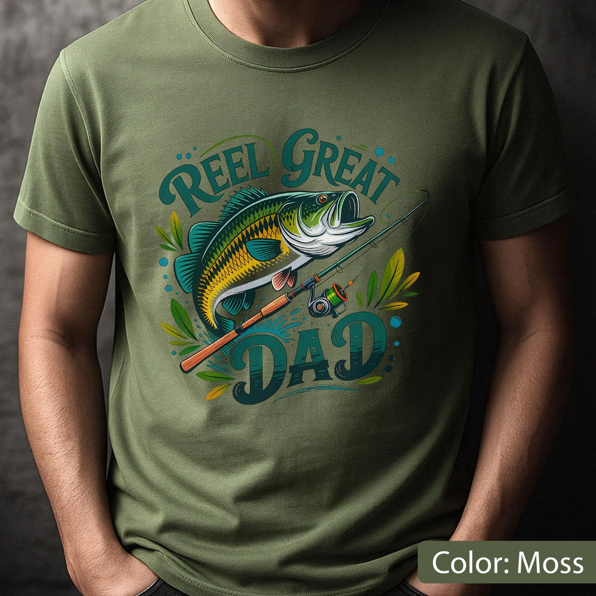 May include: Moss green t-shirt featuring a graphic of a fish, fishing rod, and the words "Reel Great Dad" in teal and yellow. The fish is green, yellow, and black. The shirt is a short-sleeved crew neck.
