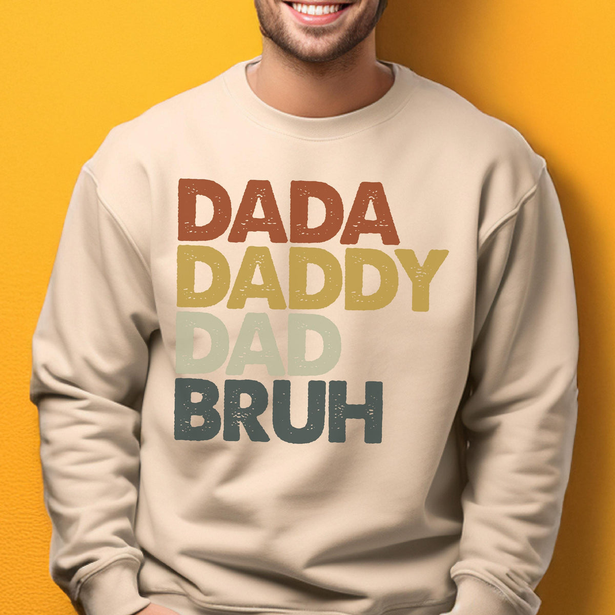 May include: A beige sweatshirt with the words "DADA DADDY DAD BRUH" printed in a stacked format. The text is in a retro color palette of brown, yellow, green, and blue. The sweatshirt is a casual style.
