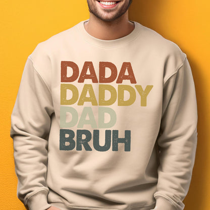 May include: A beige sweatshirt with the words "DADA DADDY DAD BRUH" printed in a stacked format. The text is in a retro color palette of brown, yellow, green, and blue. The sweatshirt is a casual style.