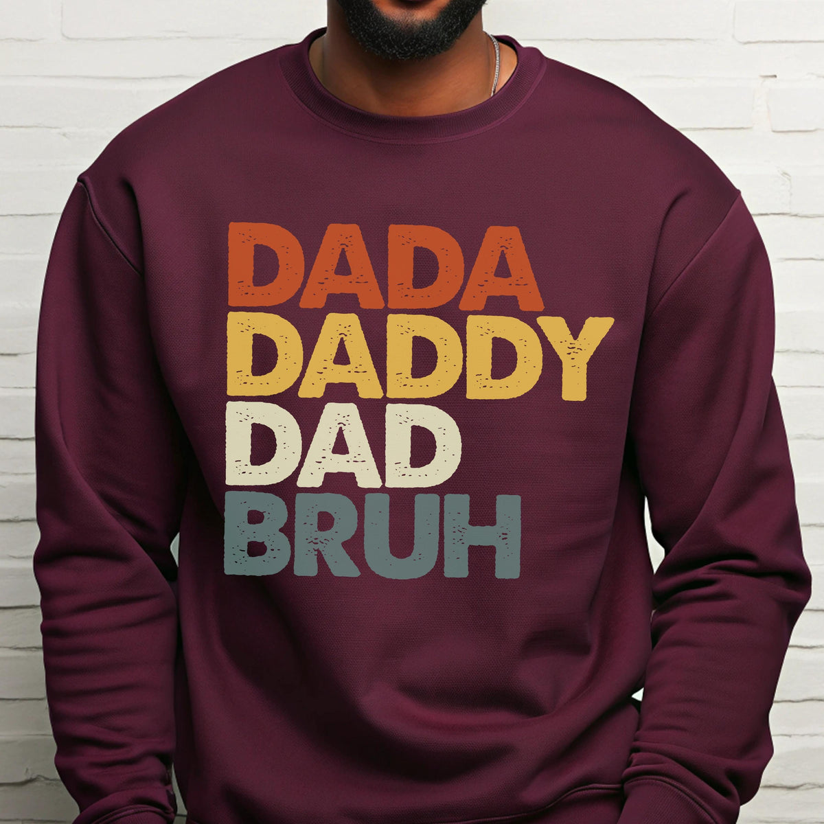 May include: Burgundy sweatshirt with the words "DADA DADDY DAD BRUH" in orange, yellow, white, and blue-green block letters. The sweatshirt has a crew neck and long sleeves.