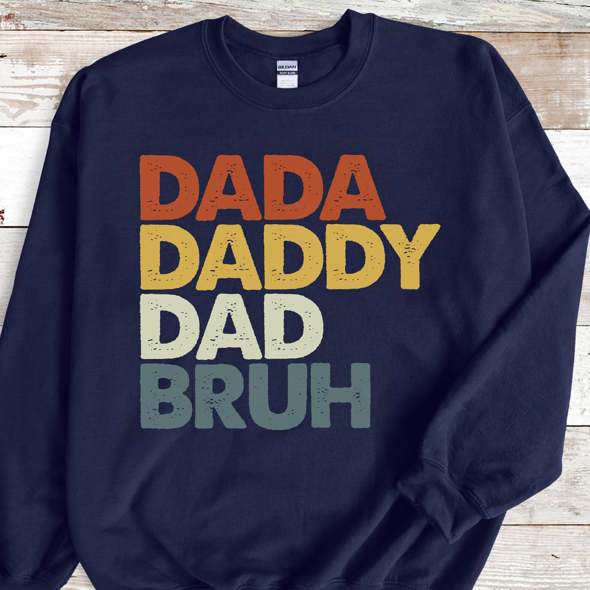 May include: Navy blue sweatshirt with the words "DADA DADDY DAD BRUH" in orange, yellow, white, and teal lettering. The sweatshirt is a casual garment, perfect for everyday wear.