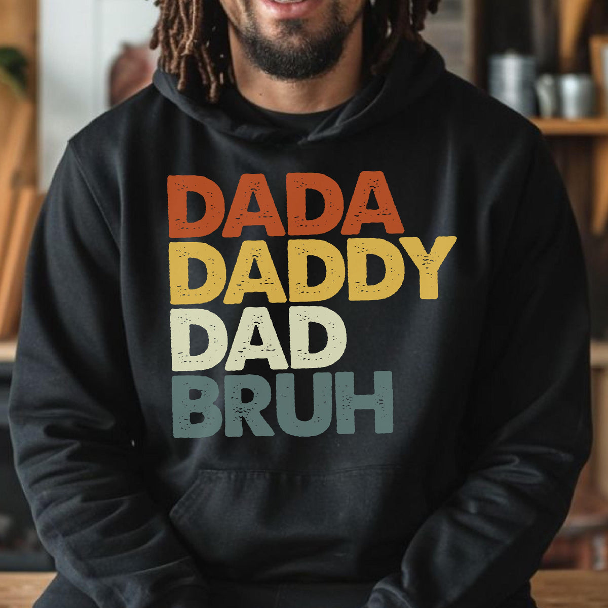 May include: Black hoodie with the words "DADA DADDY DAD BRUH" in a retro, layered font. The text colors are orange, yellow, cream, and teal. The hoodie has a front pocket and a drawstring hood.
