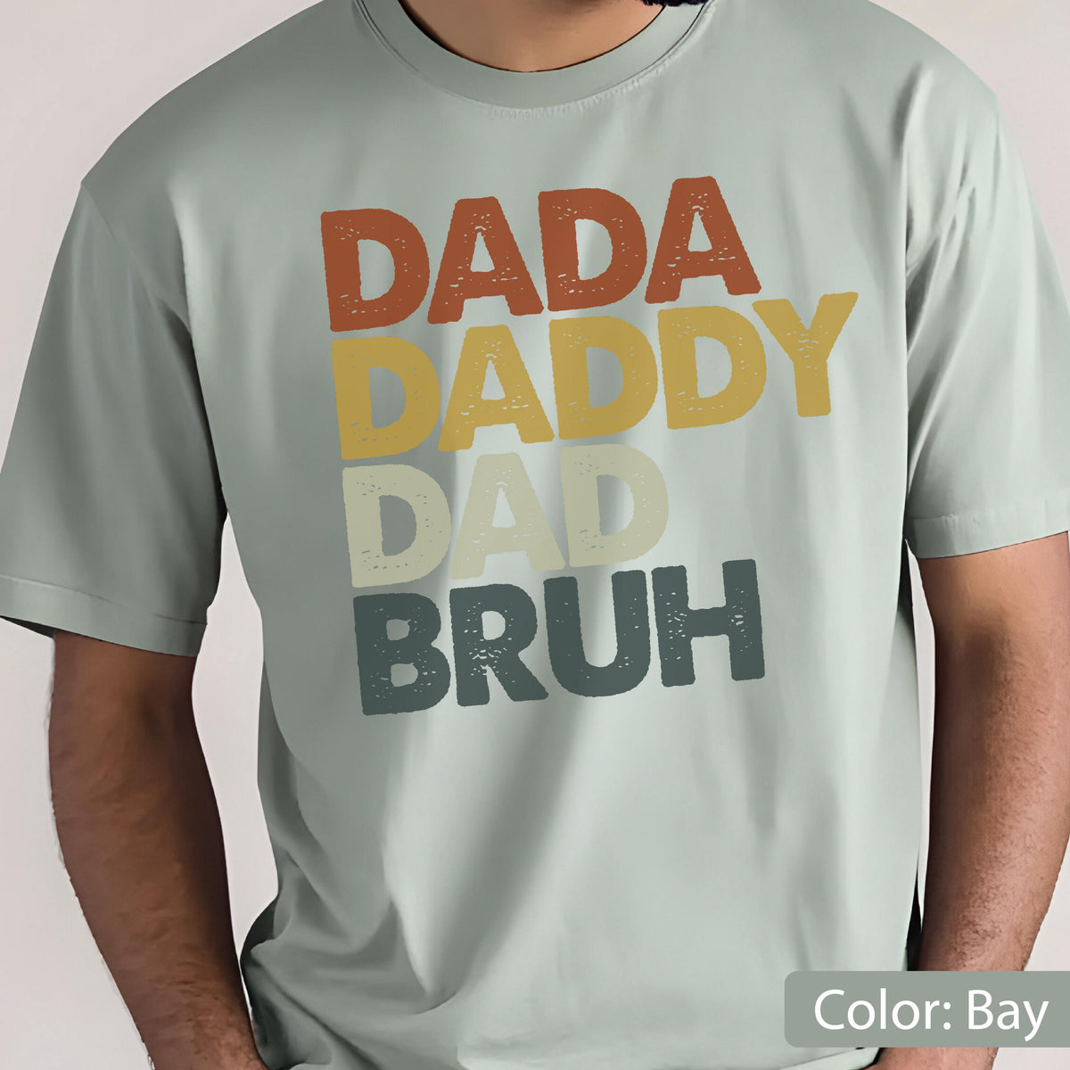 May include: A light green t-shirt with the words "DADA DADDY DAD BRUH" in a retro font. The text is in shades of brown, yellow, white, and dark green. The shirt is a solid color.