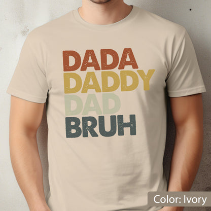 May include: Ivory-colored t-shirt with the words "DADA DADDY DAD BRUH" in a stacked, retro-style design. The text is in shades of brown, yellow, green, and blue. The shirt is a short-sleeved crew neck.