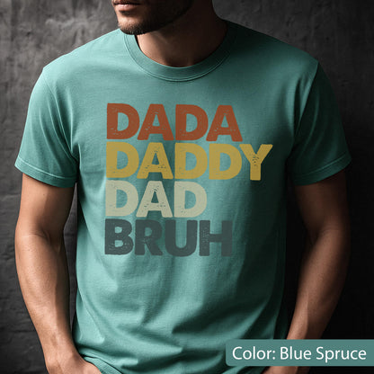 May include: Blue spruce t-shirt with the words "DADA DADDY DAD BRUH" in a retro font. The text is in shades of orange, yellow, gray, and blue. A casual tee for a dad or father figure.