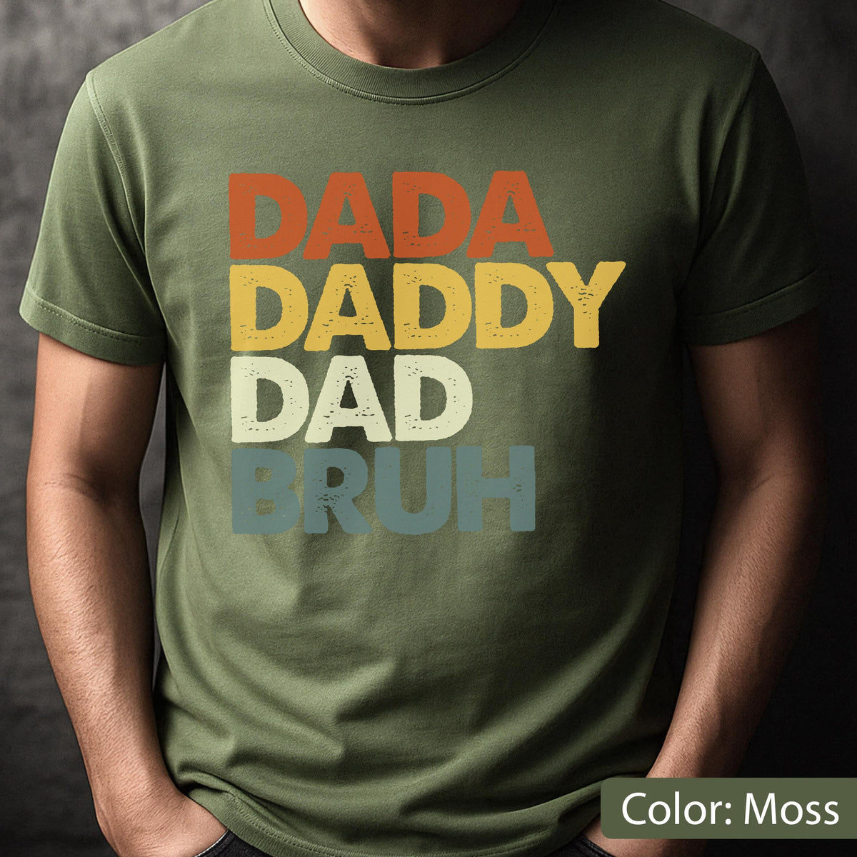 May include: Moss green t-shirt with the words "DADA DADDY DAD BRUH" in orange, yellow, white, and blue lettering. The shirt is a solid color and has a crew neck.