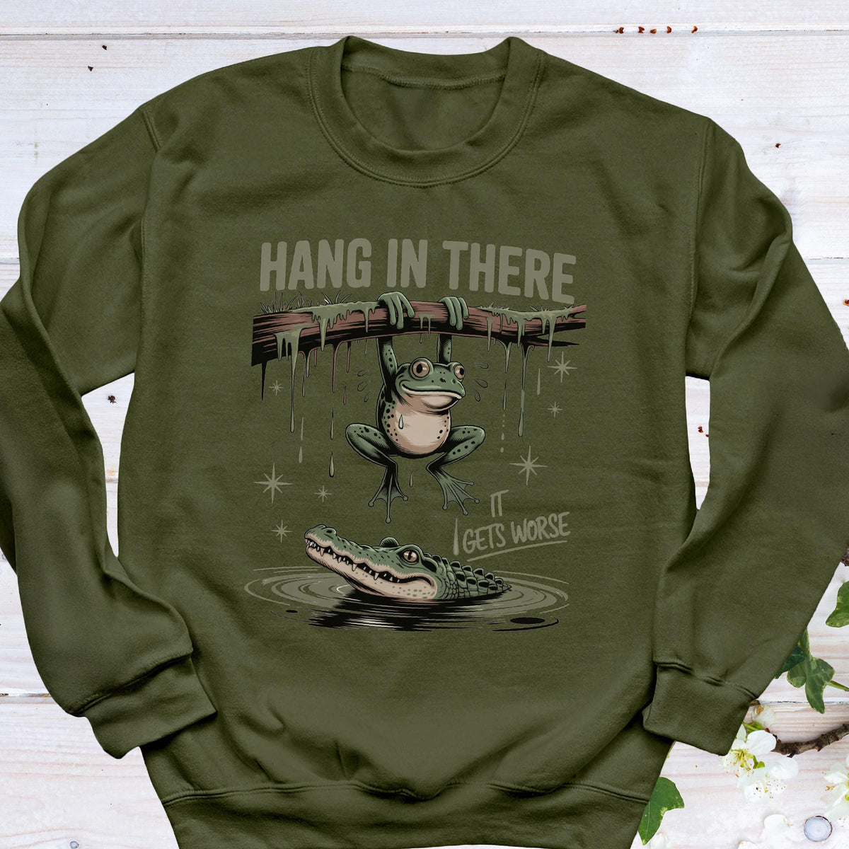 May include: Olive green sweatshirt with a humorous graphic. The design features a frog hanging from a branch above an alligator in water, with the text "HANG IN THERE" and "IT GETS WORSE".