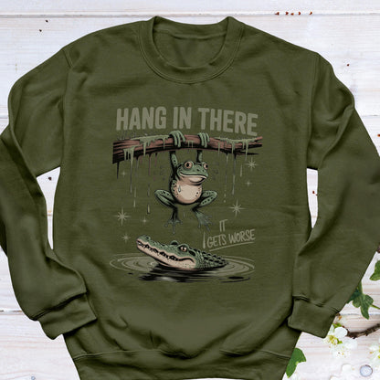 May include: Olive green sweatshirt with a humorous graphic. The design features a frog hanging from a branch above an alligator in water, with the text "HANG IN THERE" and "IT GETS WORSE".