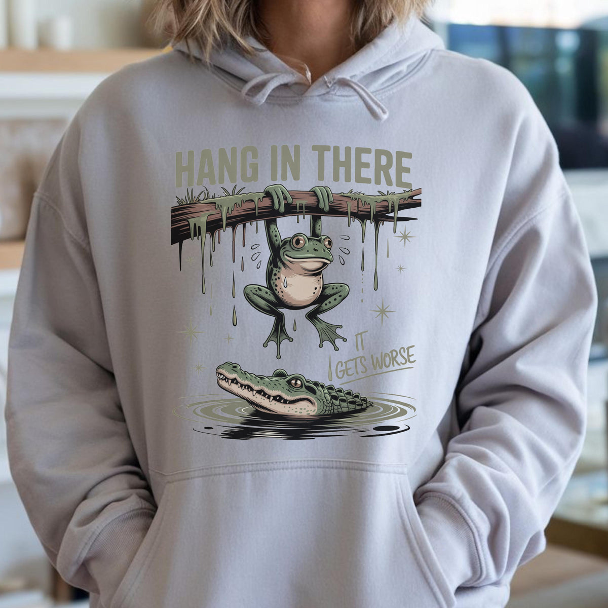 May include: A light gray hoodie featuring a humorous illustration. A frog hangs from a branch above an alligator in water. The text "HANG IN THERE" is above the frog, and "IT GETS WORSE" is below.