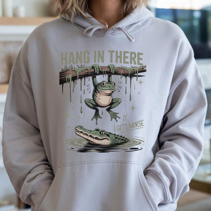 May include: A light gray hoodie featuring a humorous illustration. A frog hangs from a branch above an alligator in water. The text "HANG IN THERE" is above the frog, and "IT GETS WORSE" is below.