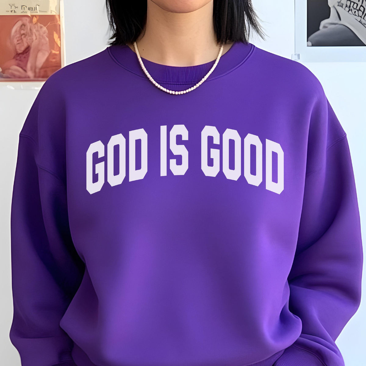 May include: A purple sweatshirt with the text "GOD IS GOOD" in white block letters. The sweatshirt has a crew neck and is worn with a pearl necklace.