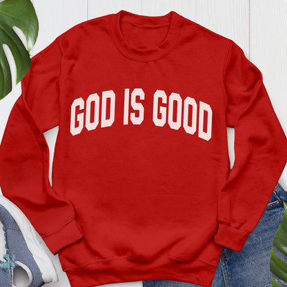 May include: A red crewneck sweatshirt with the white text "GOD IS GOOD" across the chest. The sweatshirt is made of a soft material and has long sleeves. The background includes a pair of blue jeans and a pair of blue sneakers.