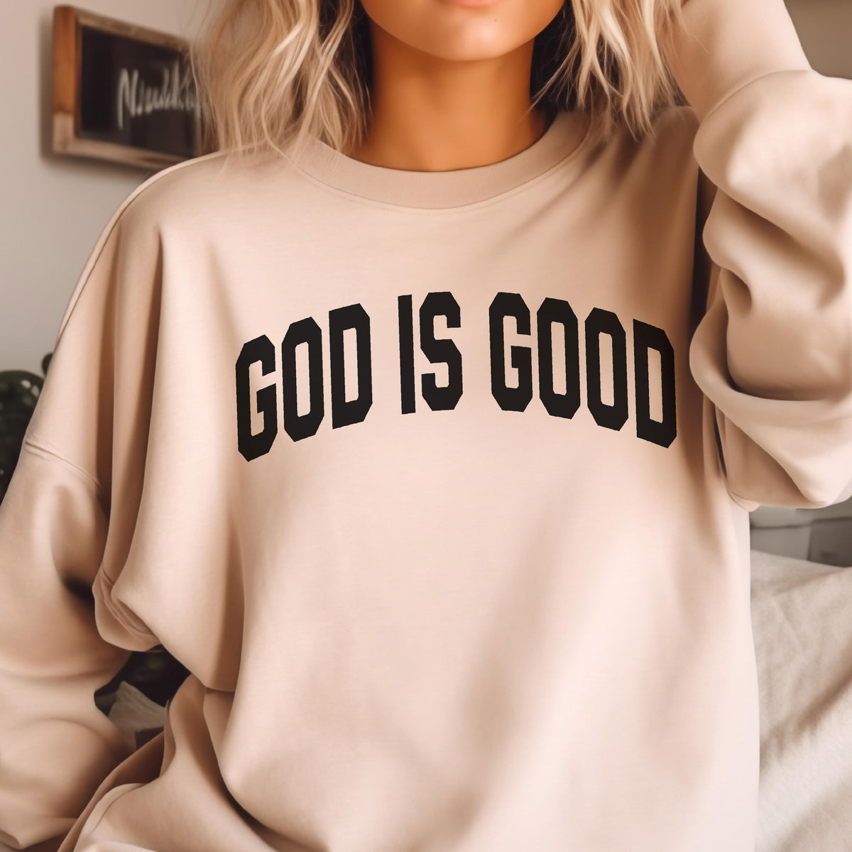 May include: A beige sweatshirt with the text "GOD IS GOOD" in bold, black letters across the chest. The sweatshirt has a crew neck and long sleeves. The background is blurred.