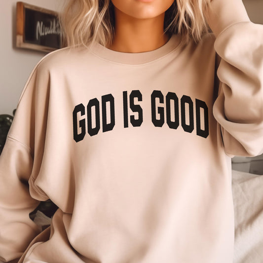 May include: A beige sweatshirt with the text "GOD IS GOOD" in bold, black letters across the chest. The sweatshirt has a crew neck and long sleeves. The background is blurred.