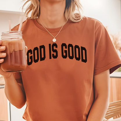 May include: A rust-colored t-shirt with the text "GOD IS GOOD" in bold black letters. The shirt has short sleeves and a crew neck. A person is holding a glass jar with a straw and a brown beverage.
