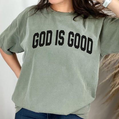 May include: Sage green t-shirt with the text "GOD IS GOOD" in bold black letters. The shirt has short sleeves and a relaxed fit. The fabric appears soft and comfortable, suitable for casual wear.