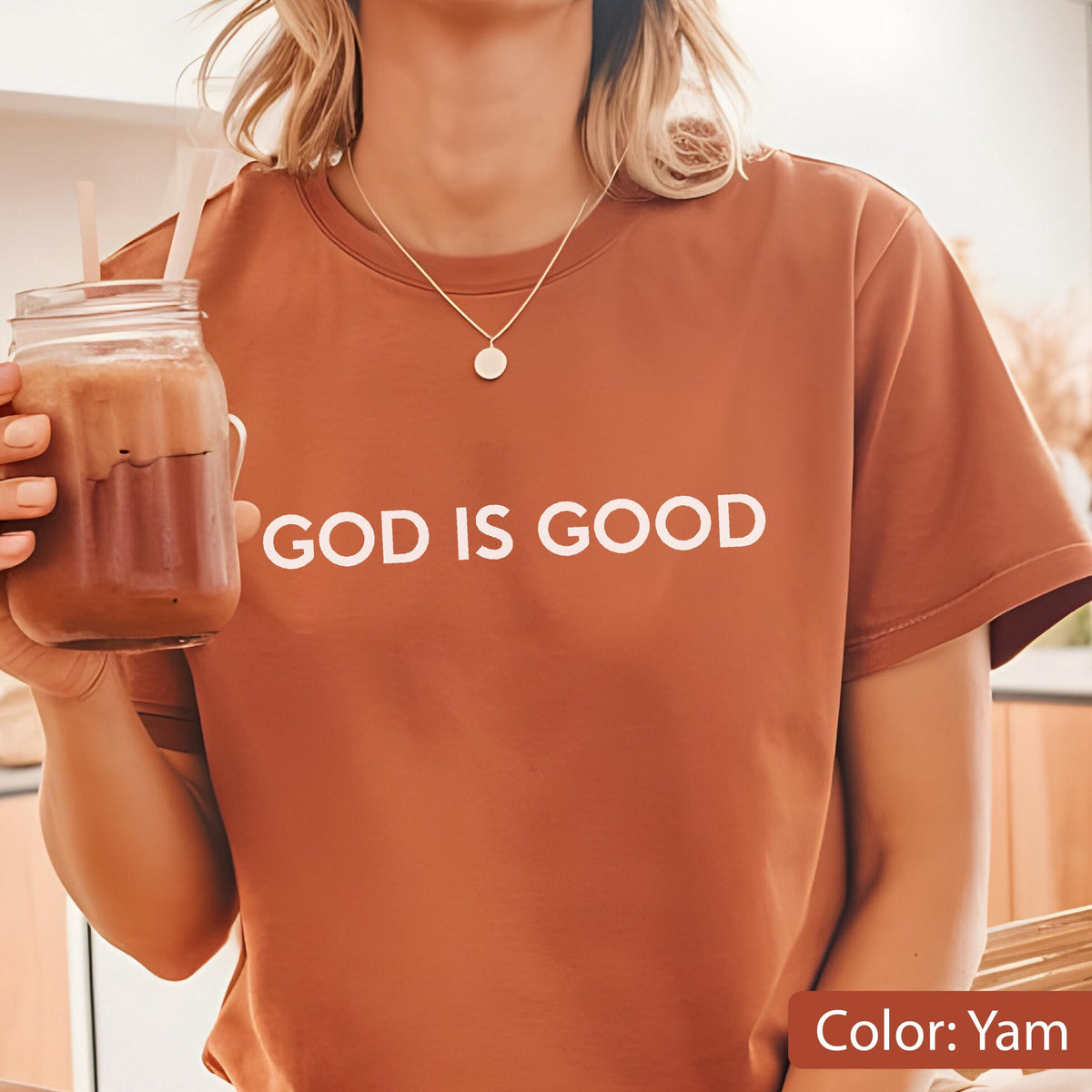May include: A short-sleeved, yam-colored t-shirt with the phrase "GOD IS GOOD" printed in white. The shirt is made of a soft material and is being worn by a person. A gold necklace with a small pendant is also visible.