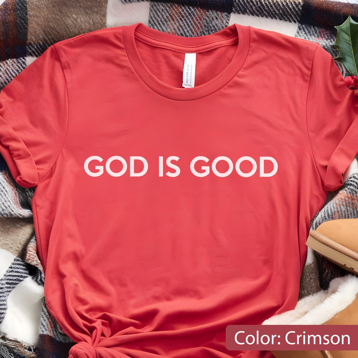 May include: Crimson-colored t-shirt with the text "GOD IS GOOD" in white block letters. The shirt is made of a soft material and has a crew neck. The shirt is folded on a plaid blanket.