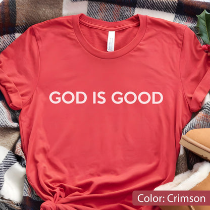 May include: Crimson-colored t-shirt with the text "GOD IS GOOD" in white block letters. The shirt is made of a soft material and has a crew neck. The shirt is folded on a plaid blanket.
