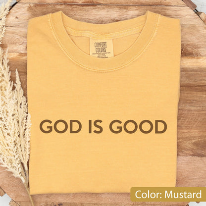 May include: Mustard-colored t-shirt with the phrase "GOD IS GOOD" printed in brown block letters. The shirt has a crew neck and short sleeves. The tag reads "Comfort Colors".