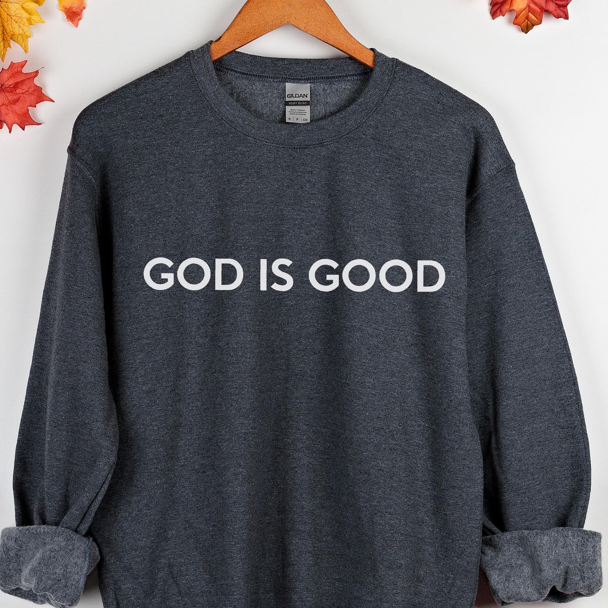 May include: Dark gray sweatshirt with the white text "GOD IS GOOD" across the chest. The sweatshirt has a crew neck and long sleeves with ribbed cuffs. The Gildan brand label is visible at the back of the neck.