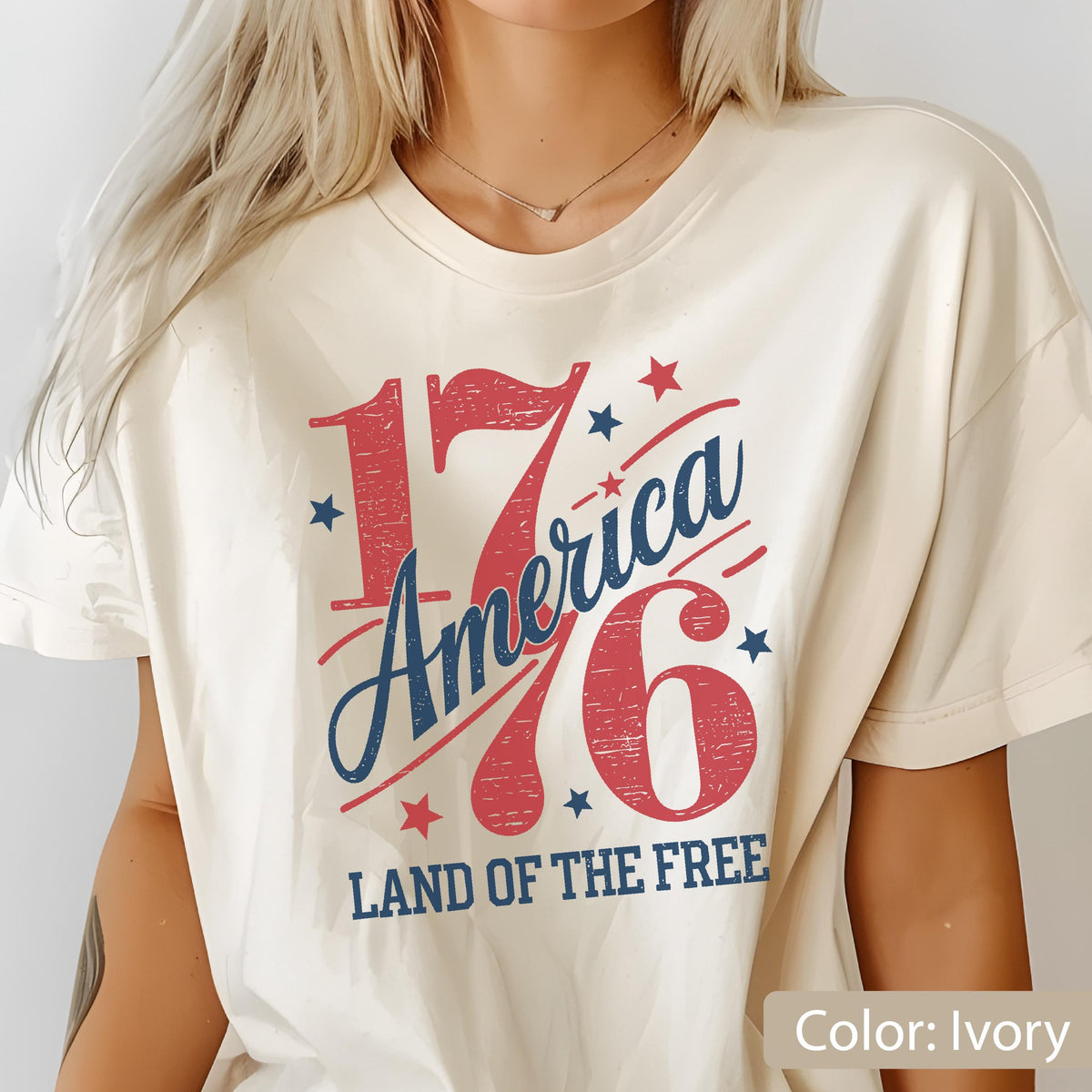 May include: Ivory-colored t-shirt with a patriotic design. The graphic features the number "176" in red, with "America" in blue script, and the text "LAND OF THE FREE." Small blue and red stars complete the design.