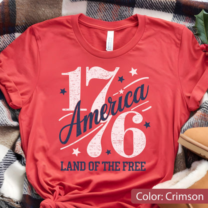 May include: Crimson red t-shirt with the text "1776 America" in white and navy blue, along with the words "Land of the Free" in white. The shirt is folded, and the color is noted as crimson.