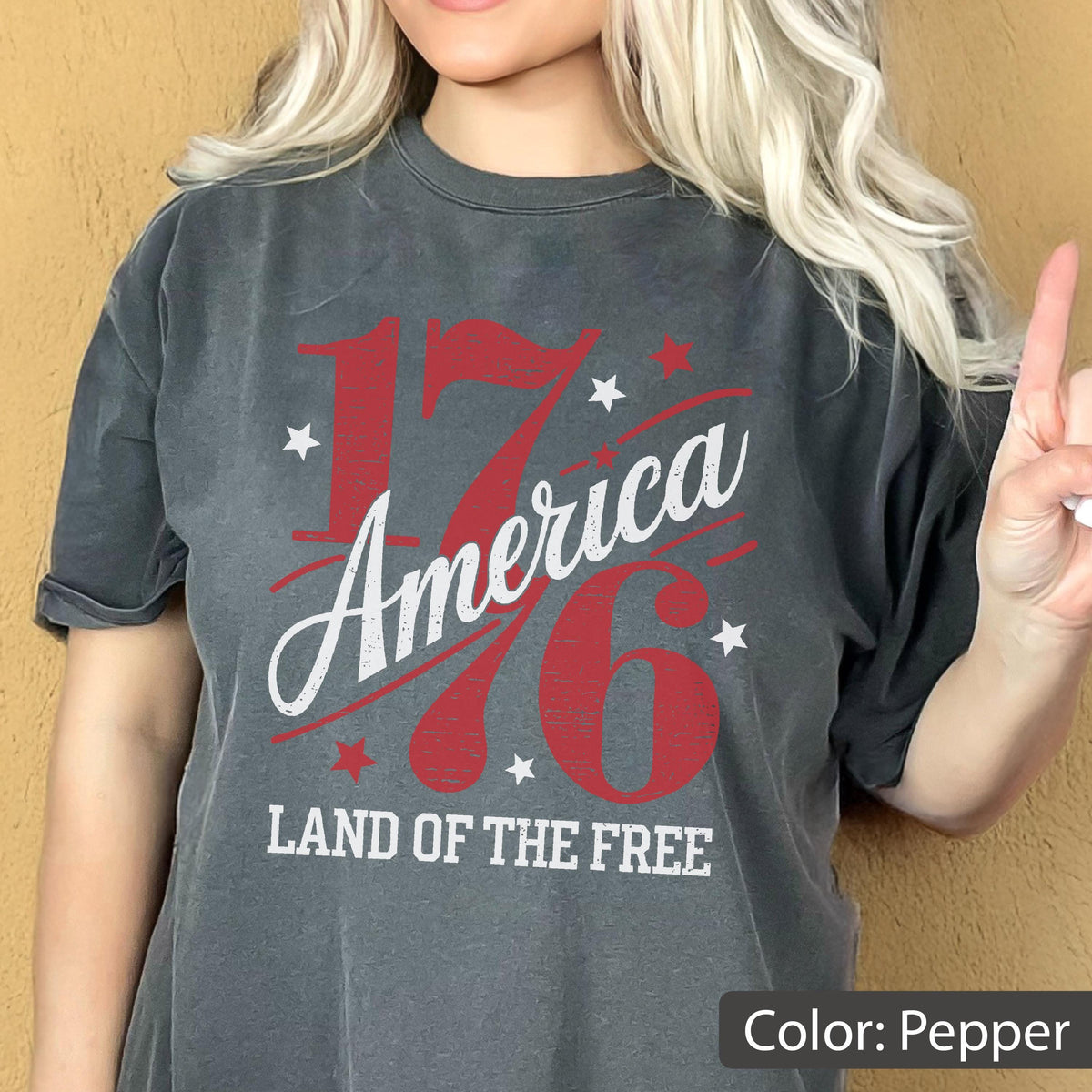 May include: Pepper-colored t-shirt with the text "1776 America" in red and white, along with star graphics. The phrase "LAND OF THE FREE" is printed below. The shirt has a vintage, worn look.