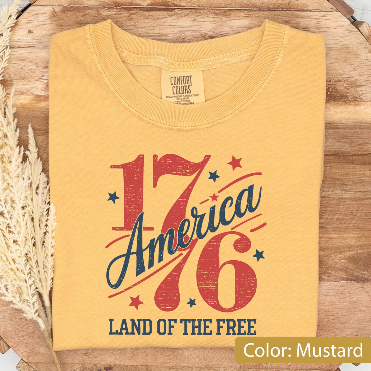May include: Mustard-colored t-shirt with the text "1776 America" in red and blue, along with stars and the phrase "Land of the Free." The shirt is made of a soft material and is folded neatly.