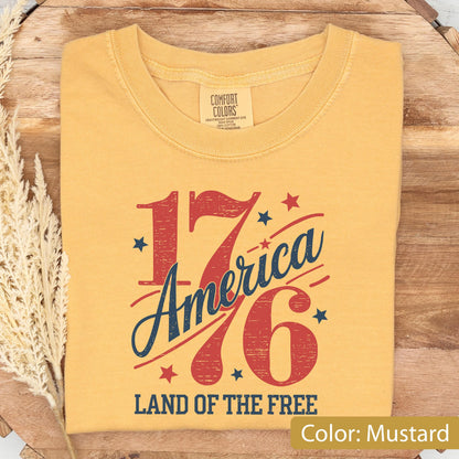 May include: Mustard-colored t-shirt with the text "1776 America" in red and blue, along with stars and the phrase "Land of the Free." The shirt is made of a soft material and is folded neatly.